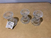 Top angled view showing three clear crystal candle holders arranged in a row, with vertical ridges and circular bases visible. One holder has a tag reading 'Athena's Fine'.