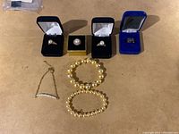 Six pieces of jewelry laid out on surface: three rings in boxes, two pearl bead bracelets, one small pearl and chain bracelet.