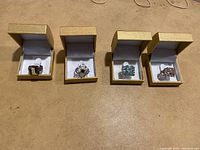 Four rings each placed inside separate gold-tone small boxes with white interior on a flat surface, showing variety of styles including gemstone and floral themes.