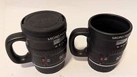 Two black ceramic espresso mugs designed to look like camera lenses, showing detailed lens markings and one with a black rubber lid on top.