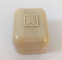 Closed cream bakelite engagement ring box showing rounded edges and raised floral design on top with ribbed detail.