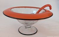 Side angle showing the clear glass footed bowl with its wide orange rim edged in black and the accompanying curved serving spoon inside the bowl.