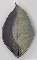 Photo of a ceramic ornamental leaf plate with detailed vein textures, dual-tone glaze half dark purple and half muted greenish-gray, representative of a decorative serveware item.