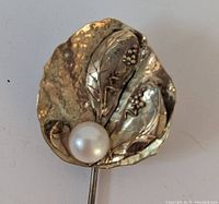 Close-up photo of gold colored brooch pin showing detailed textured leaf design with vine and berry motifs and a white pearl at the base.