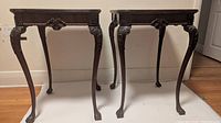 Front view of two dark wood side tables with ornamental legs and carved details on apron and legs.