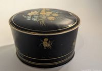 Oval black metal candy tin with painted floral design on the lid and sides, showing minor wear and a small dent on the lid.