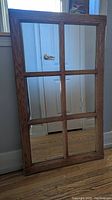 Photo of a rectangular six-panel wooden framed mirror leaning against a wall on wood flooring.