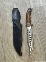 Full view of knife and black leather sheath on light wood floor. Knife blade has hole pattern, wood-grain style handle with animal head pommel.
