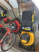 Photo of a yellow and black DEWALT cordless lawn mower with no batteries or charger visible, placed beside a red bicycle and other items on a concrete floor.