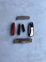 Photo of five folding cutting tools arranged on a grey surface, showing various sizes, handle materials including metal, wood, and plastic, and including a multi-tool with corkscrew.