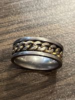 Close-up photo of the titanium ring with gold-colored metal chain inlay on a wooden surface, showing side angle of the ring.