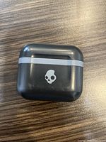 Closed black Skullcandy charging case with skull logo on front, sitting on wooden surface.