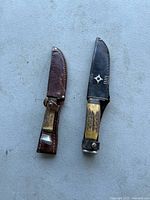Two vintage hunting knives with stag handles and leather sheaths on light background. One sheath decorated with silver-tone star and lines.