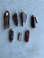 Top view of seven vintage folding cutters on a grey surface with varied shapes and handles
