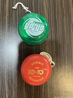 Photo showing two yo-yos on a dark wood surface: a green translucent 'Premium yo-yo' and a red 'Authentic Pro Yo-yo Tonfuque Authentique'. Both have strings attached and appear in good condition.