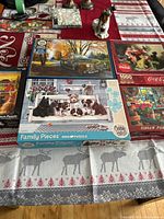 Five boxed puzzles displayed on a red and white tablecloth with winter and Christmas motifs. The puzzles feature a variety of themes such as dogs, vintage trucks, a Coca-Cola Christmas image, and a Christmas shop scene.