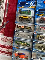 Partial view of unopened early Hot Wheels cars in original blue Mattel packaging showing various models including a green car, yellow car, orange hot rod, and others.