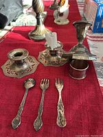 Photo of 3 candle holders, 2 forks, 1 spoon, and 1 napkin ring arranged on a red tablecloth.