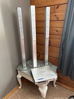 Four silver tower speakers positioned beside each other, placed on a small white table against a wooden wall and a curtain in the background.