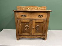 Front view of a small antique wooden sideboard with drawer and two cabinet doors, decorative hardware and carved details.