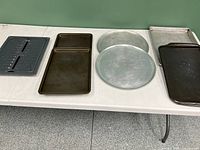 All items grouped together showing four rectangular baking trays, two round baking trays, and one rectangular grease tray on white surface with grey floor.