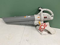 Full view of Ryobi electric leaf blower and mulching vacuum with large gray nozzle and attached collection bag on floor against green wall.