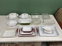 Full lot overview of glass and ceramic baking and casserole dishes on table against green wall.