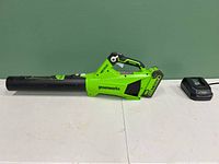 Full side view of Greenworks cordless electric leafblower with battery attached and charger unit beside it on a white surface against a green wall.