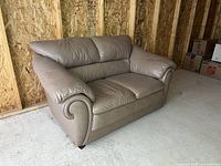 Beige faux-leather loveseat angled side view showing curved padded armrest and backrest cushion on an unfinished wood floor with wooden wall paneling behind.