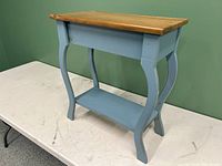 Small wooden end table with natural wood top and painted blue sides and shelf from front angled view.