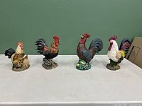 Four small standalone painted ceramic rooster figurines in various natural colors and sizes up to 11 inches tall displayed on a flat surface.