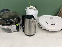 Overall shot showing T-Fal ActiFry air fryer, George Foreman grill, Betty Crocker rice cooker, and stainless steel electric kettle on a table against green background.