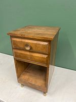 View of wooden side table front and right side showing drawer with one knob, missing knob on left, open bottom shelf, brass-capped feet.