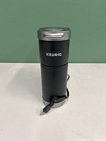 Front view of compact black Keurig coffee machine with silver top and power cord wrapped around base