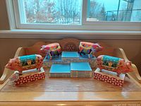 Four colorful pig figurines by Jim Shore with two matching stands on wooden surface in front of window.