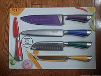 Top-down image of four knives and one peeler each with colored handle and matching blade guard, on packaging board.