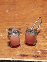 Front and side angle view of the pair of orange moonstone rectangular cabochon earrings set in silver and platinum lever back mountings, resting on wooden surface.