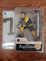 Front view of the Phil Esposito figure inside its clear plastic packaging, showing figure and included hockey stick accessory.