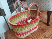Full view of hand woven Ghana basket with vibrant red and green pattern and leather handles on hardwood floor.