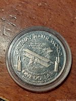 Close-up image of the commemorative coin showing the airplane design and inscriptions, enclosed in protective capsule on wooden surface.