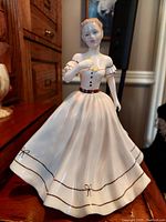 Front and slightly angled view of the Coalport 'Geraldine' vintage porcelain figurine showcasing the white gown with brown trim and hand-painted details, set on a wooden surface.