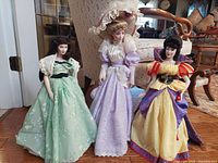 Three porcelain dolls arranged in living room setting. Snow White doll in yellow dress, tall doll in lavender Victorian dress, and doll in green floral dress with shawl.