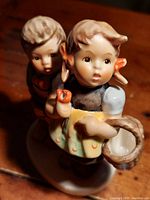 Close front left angle view of vintage Hummel porcelain figurine showing two children on an oval base, girl holding orange and basket, boy standing beside her.