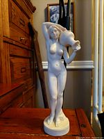 Front view of vintage white matte sculpture of nude female holding water jug on shoulder.
