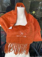 Orange alpaca wool scarf displayed draped over chair showing fringe and branded embroidery.