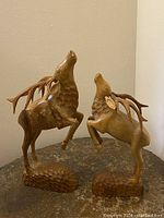 Photo showing two hand carved wood elk sculptures on a brown surface against a beige wall, highlighting the full figures and pedestal bases.