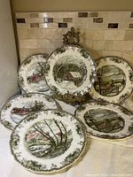 Six Johnson Bros The Friendly Village pattern dinner plates arranged with one on stand and five laid out, showing different hand-colored rural village scenes with green grapevine decorated rims.