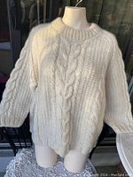 Front view of large cable knit sweater showing off-white wool material and thick cable knit pattern with ribbed collar, cuffs and hem.