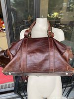 Front view of brown full grain Italian leather duffel bag displayed on mannequin torso. Shows rich leather texture and metal feet on base.