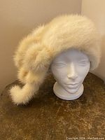 White mink hat showing front angle on a mannequin head with decorative fur element on side and trailing fur.